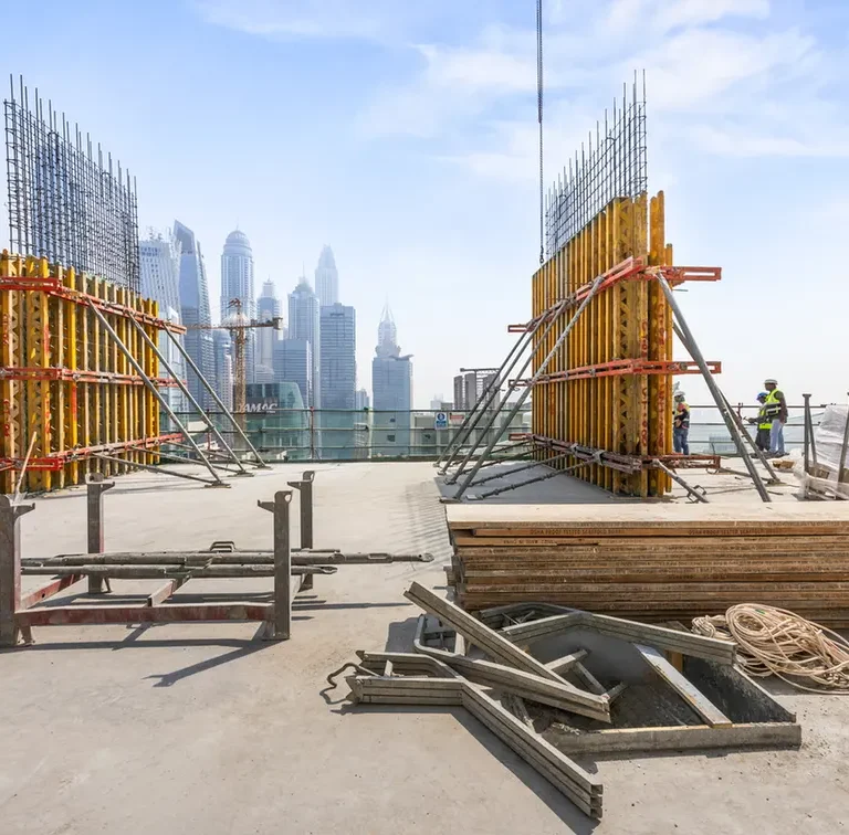 Construction site on a rooftop with wooden and metal formwork, rebar, and workers; city skyline in the distance.