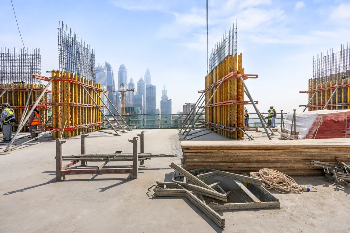 Construction site on a rooftop with wooden and metal formwork, rebar, and workers; city skyline in the distance.