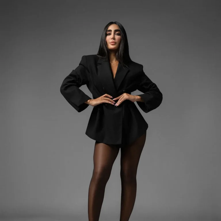 Full-body portrait of a woman in a black blazer dress, hands on hips, against a gray studio backdrop.