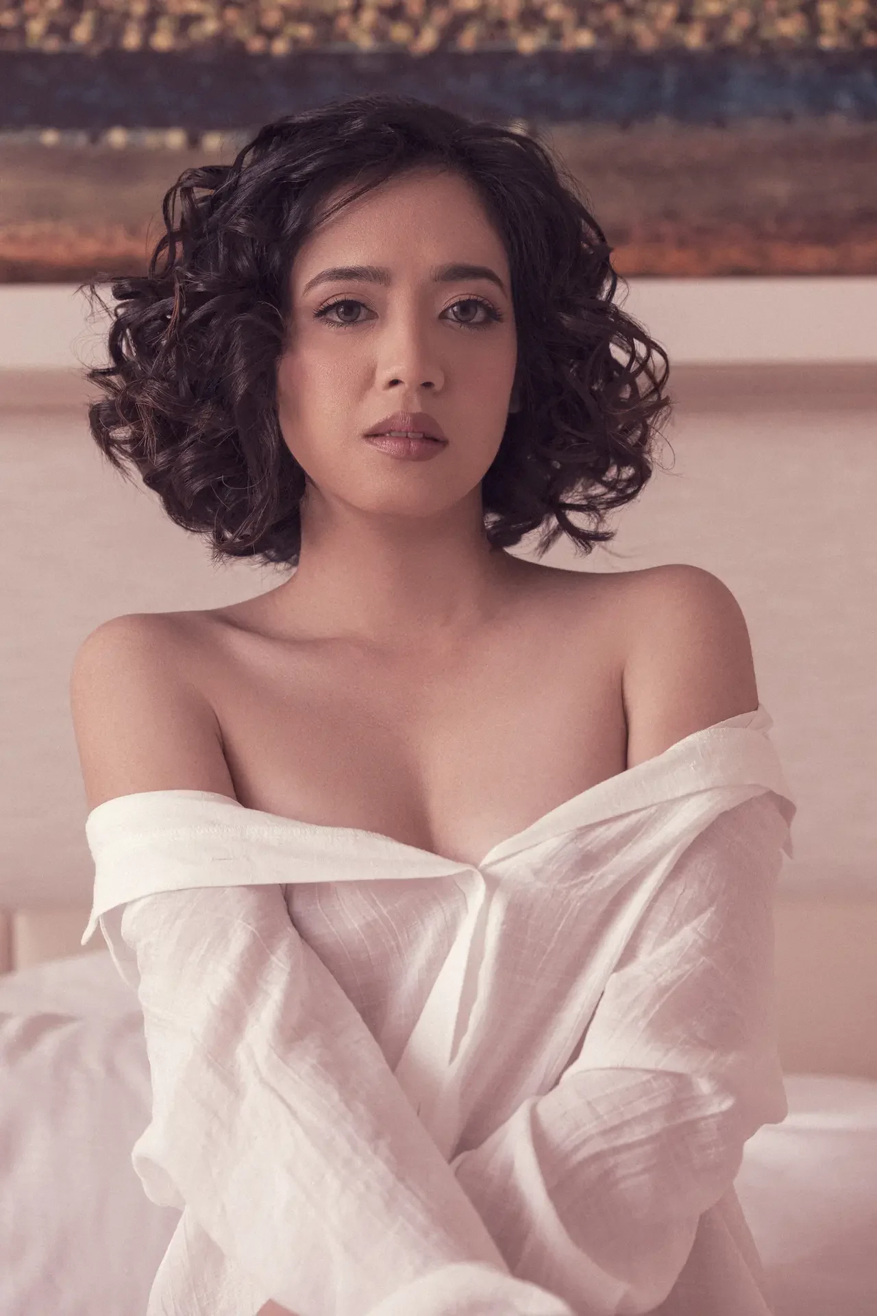 Portrait of a woman with short curly dark hair, wearing a white off-the-shoulder blouse, looking softly at the camera.