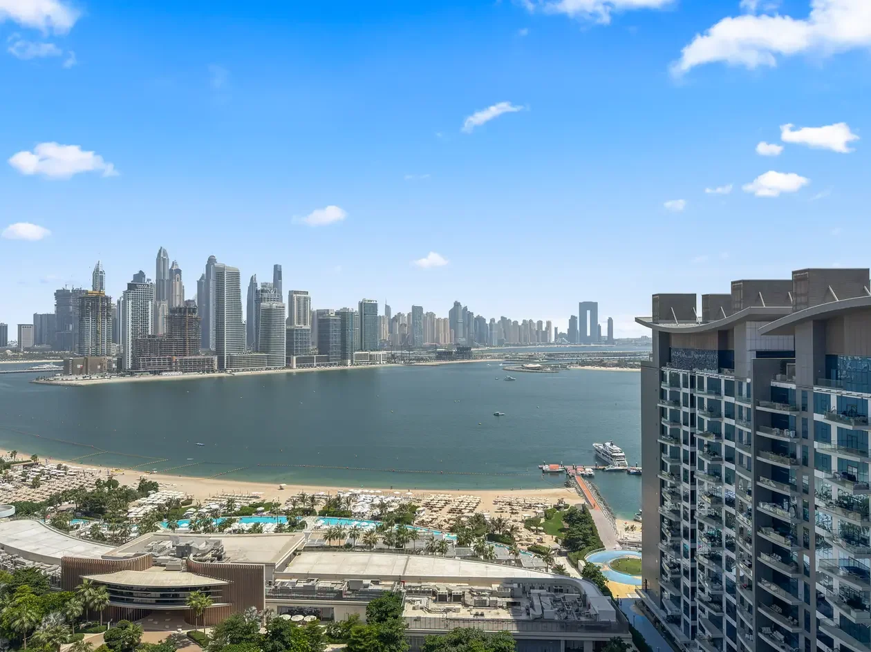City skyline along a calm bay with high-rise buildings and a sandy beach in the foreground, under a bright blue sky.