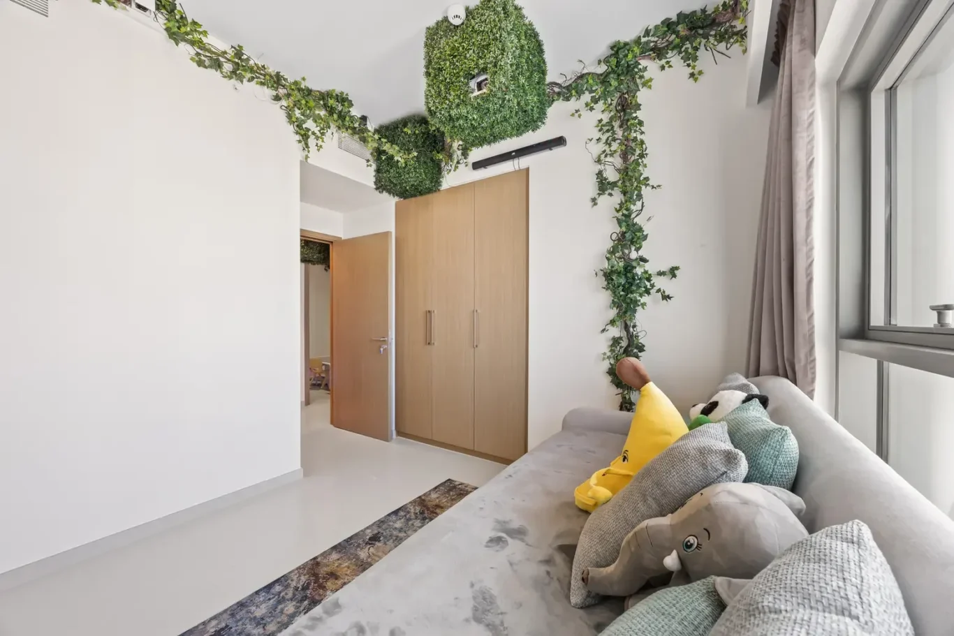 Living room with a gray sofa and stuffed toys, large window with gray curtains, and a wooden built-in wardrobe; ivy-adorned ceiling and walls add greenery.