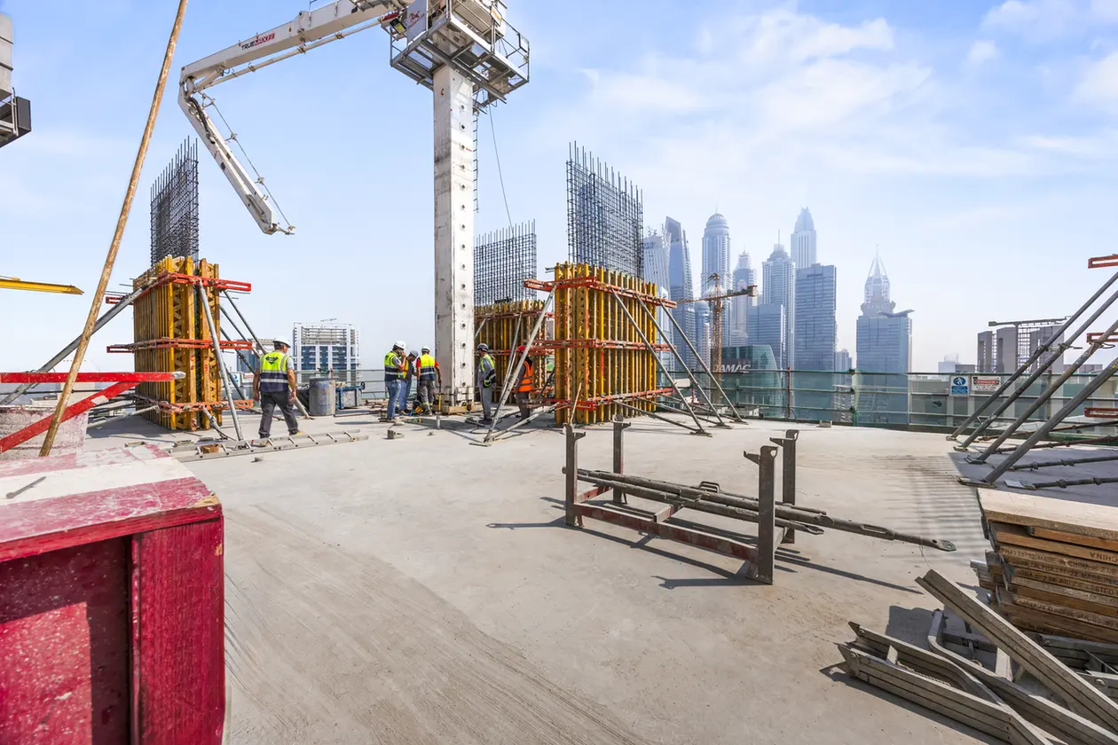 Construction workers in safety gear stand around a concrete column on a rooftop with a city skyline in the background,