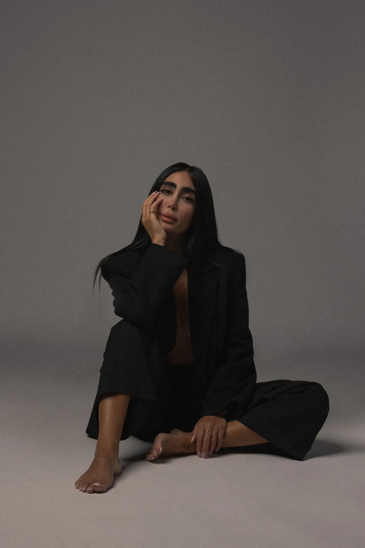 Woman with long dark hair sits on the floor in a black blazer, against a gray studio backdrop, resting her face on her hand.