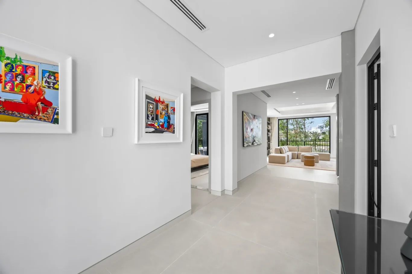 White hallway with two framed colorful art pieces, leading to a bright living area with beige sectional and large windows.