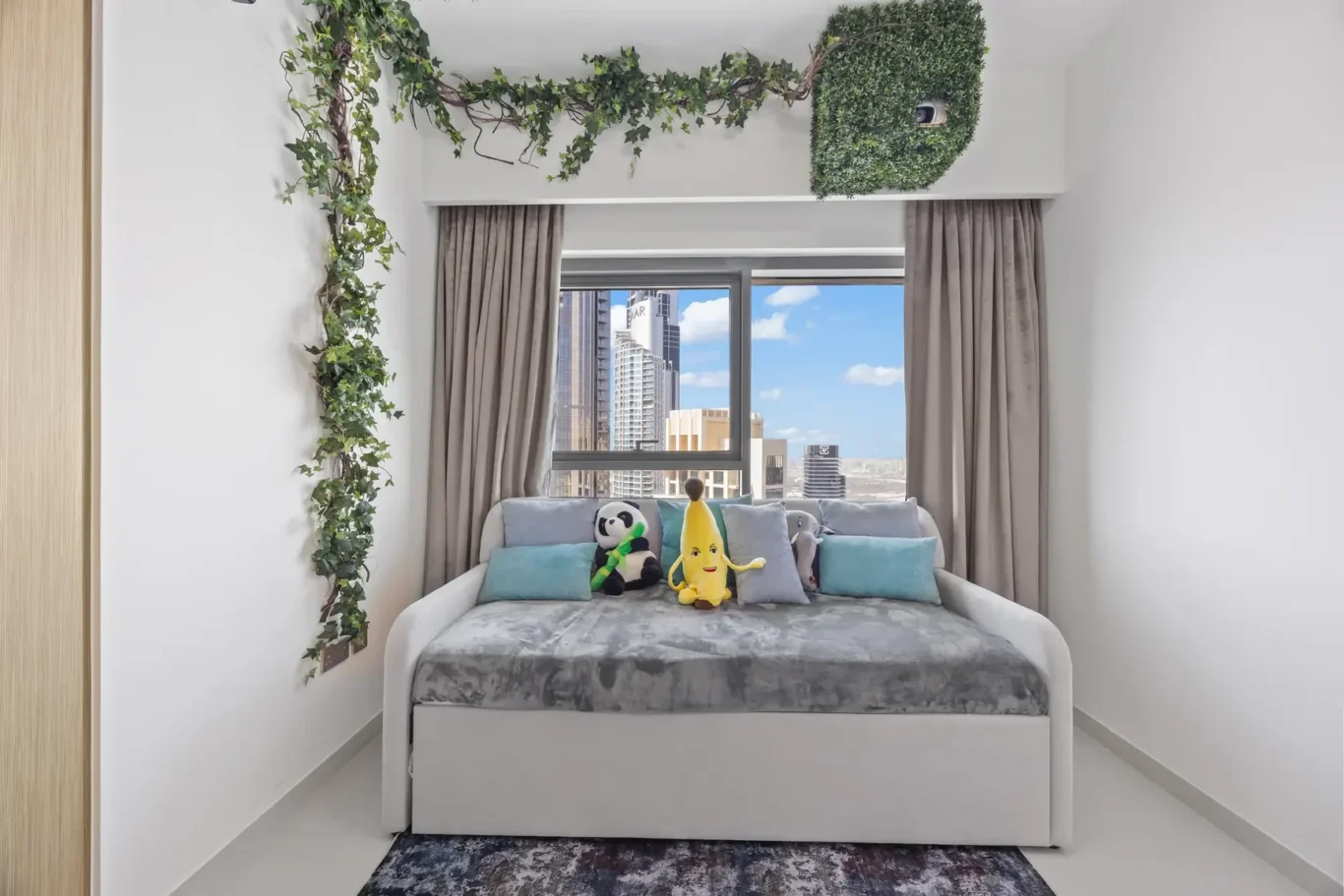 White-walled bedroom with beige curtains and a large window showing a city skyline; a bed with gray blanket and blue cushions, plus panda, banana, and elephant plush toys on top.