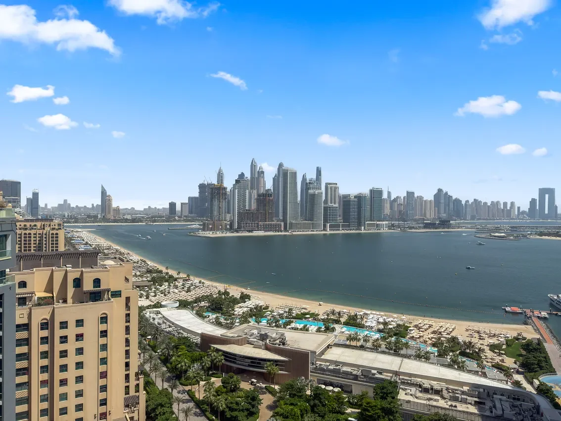 Panoramic view of a coastal city skyline across a calm bay, with high-rise buildings and a sandy beach in the foreground under a blue sky