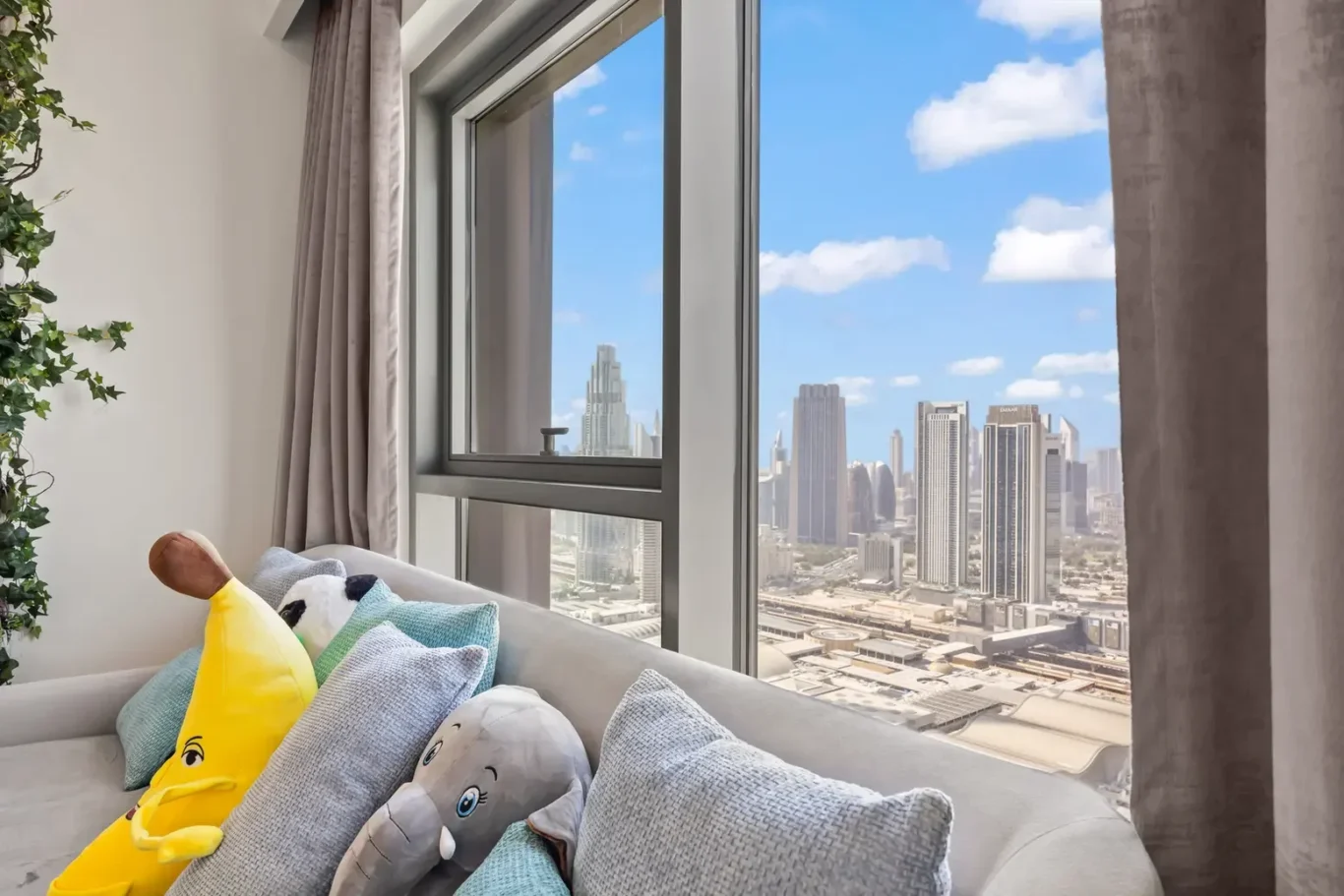Cozy living room corner with a large window showing a city skyline and blue sky; gray sofa with light blue cushions and stuffed yellow banana toy nearby.