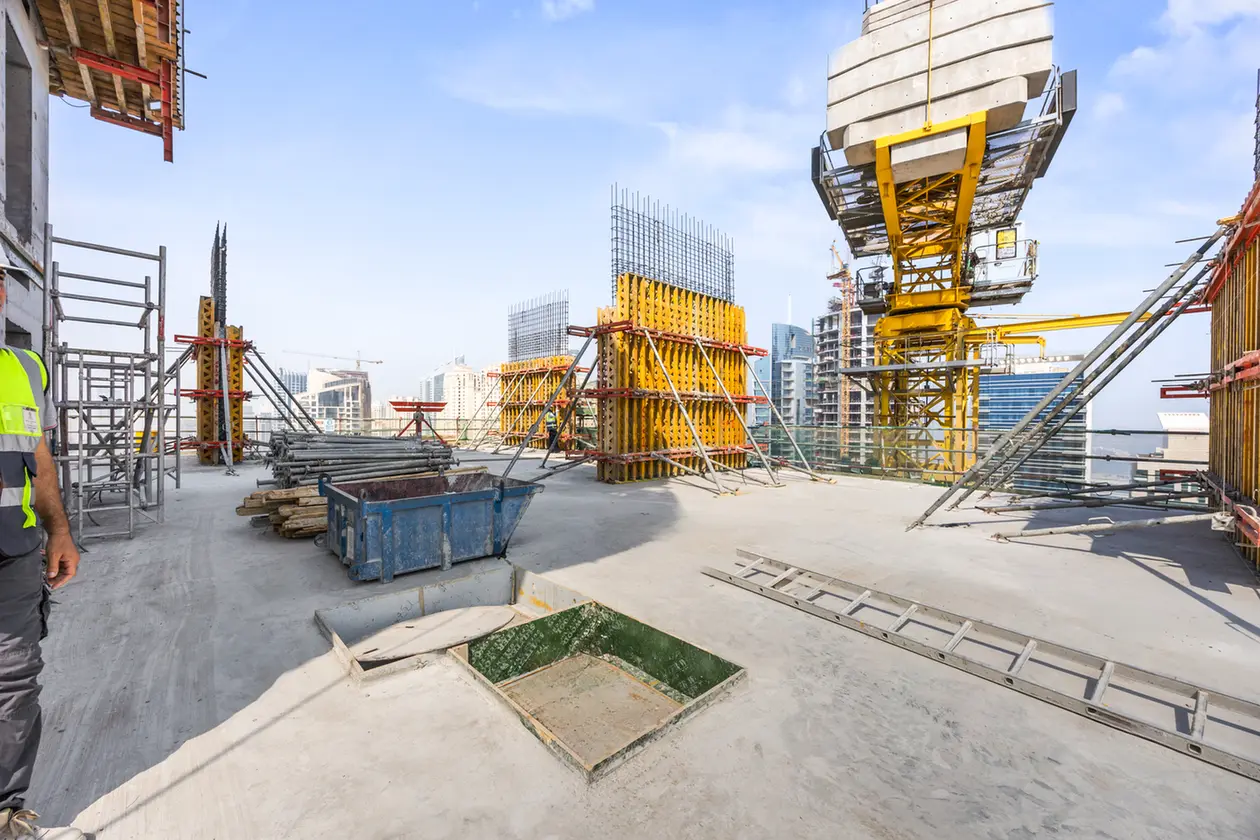 Construction site on a high-rise floor with yellow formwork, scaffolding, and steel rods against a city skyline.