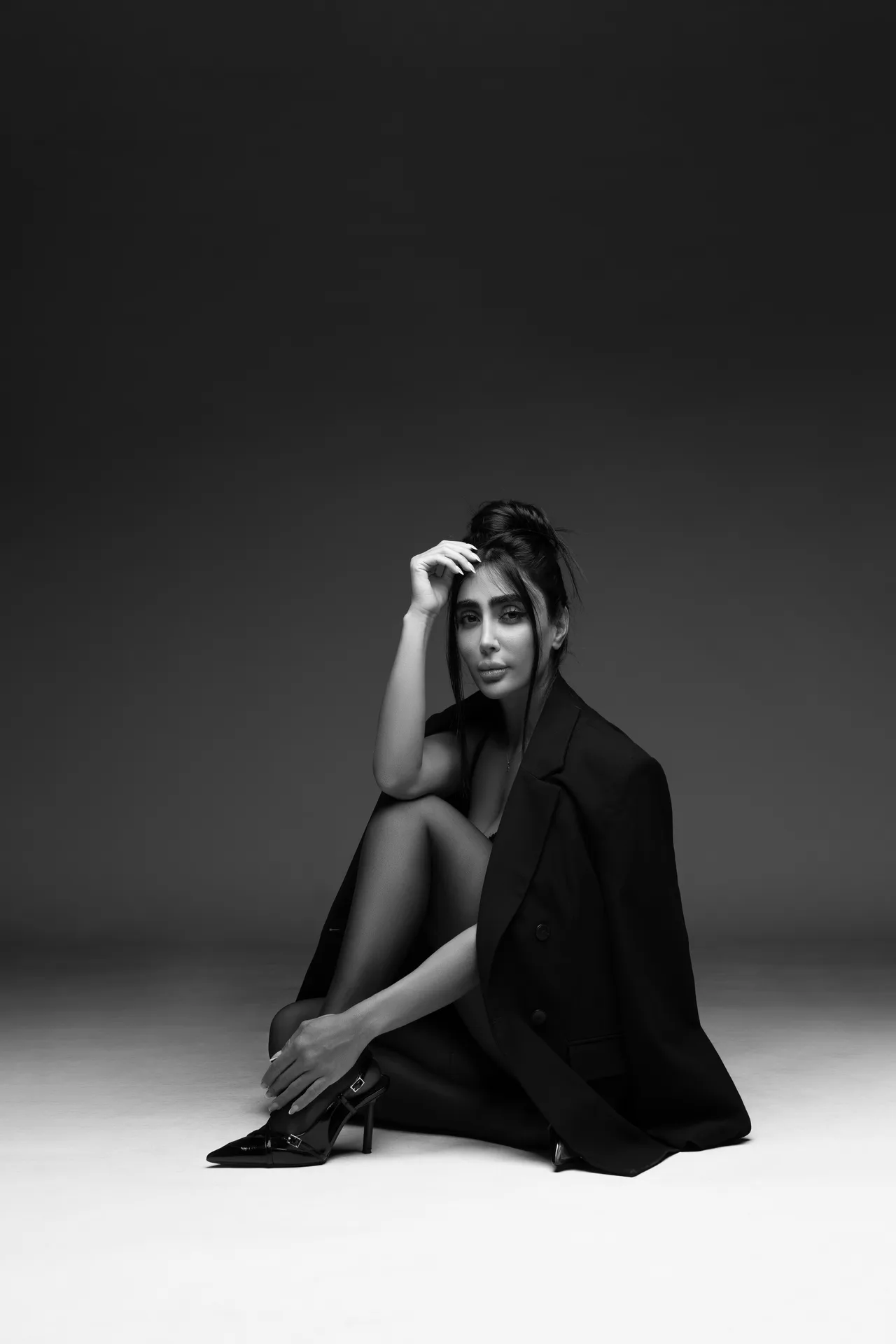 Black-and-white portrait of a woman seated on the floor, wearing a blazer and high-heeled shoes, hand resting on her hair.