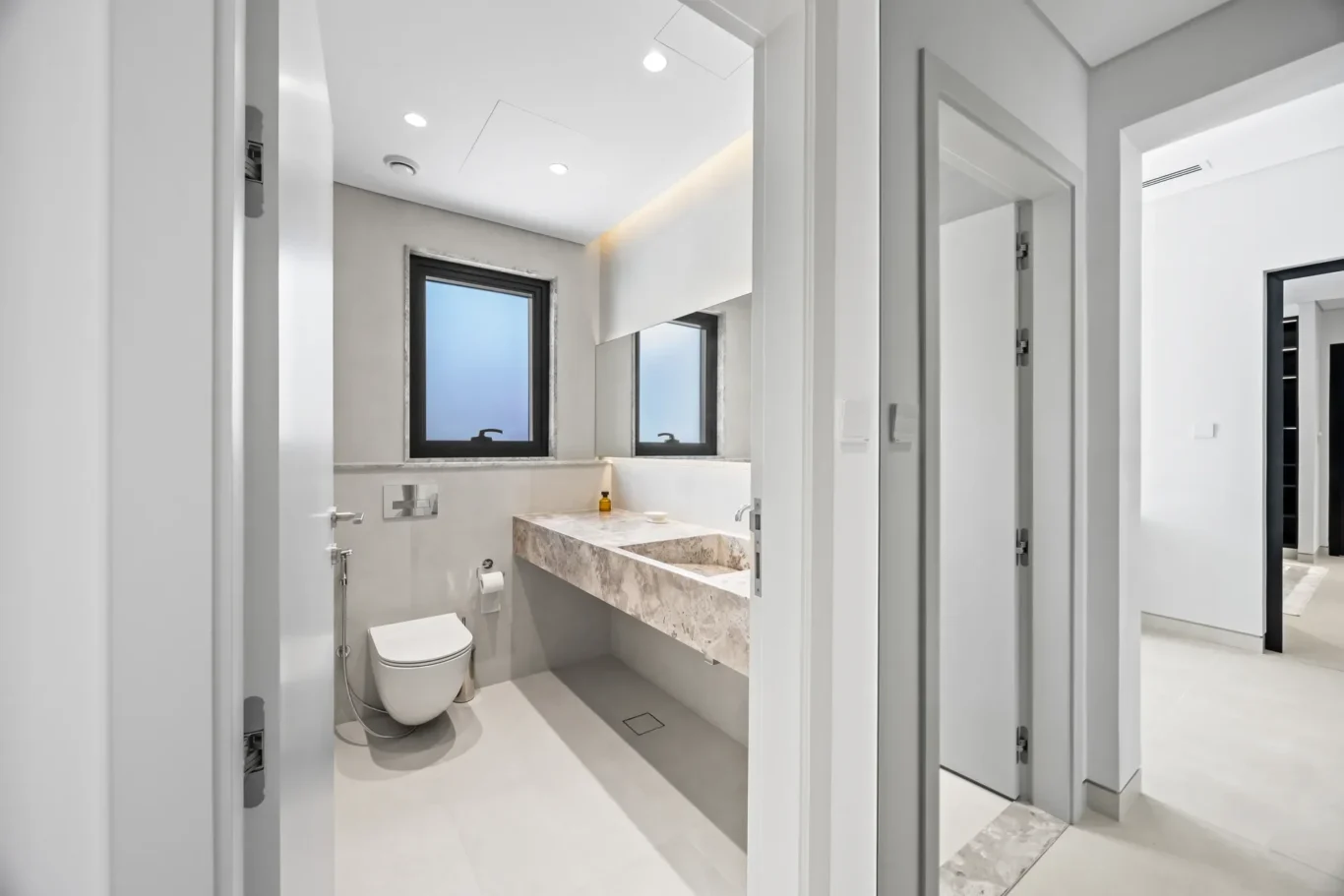 Modern white bathroom with a double marble countertop, wall-mounted toilet, and two black-framed windows viewed from the doorway.