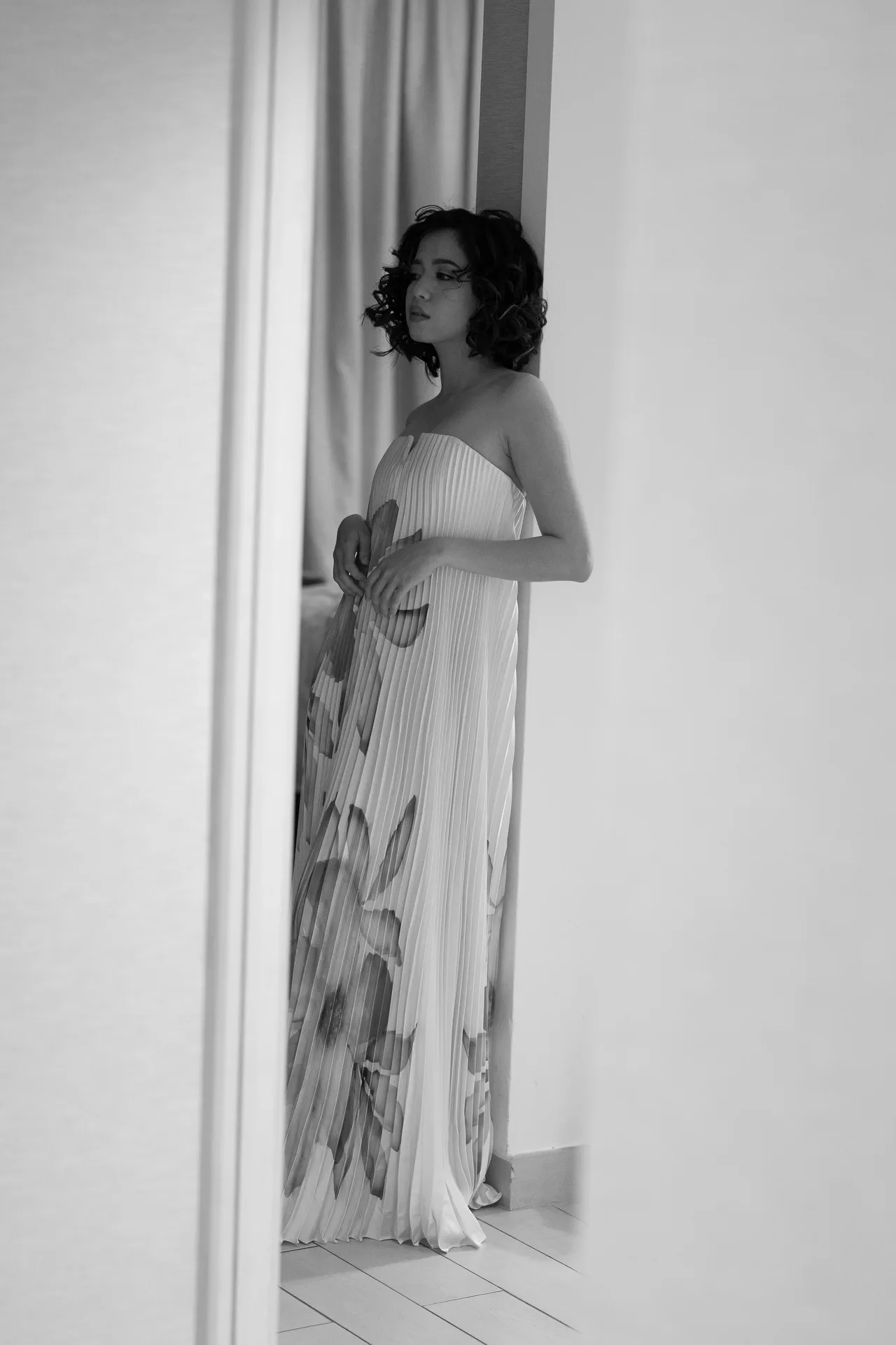 A woman in a strapless, pleated dress stands by a doorway, looking to the left with hands near her waist."