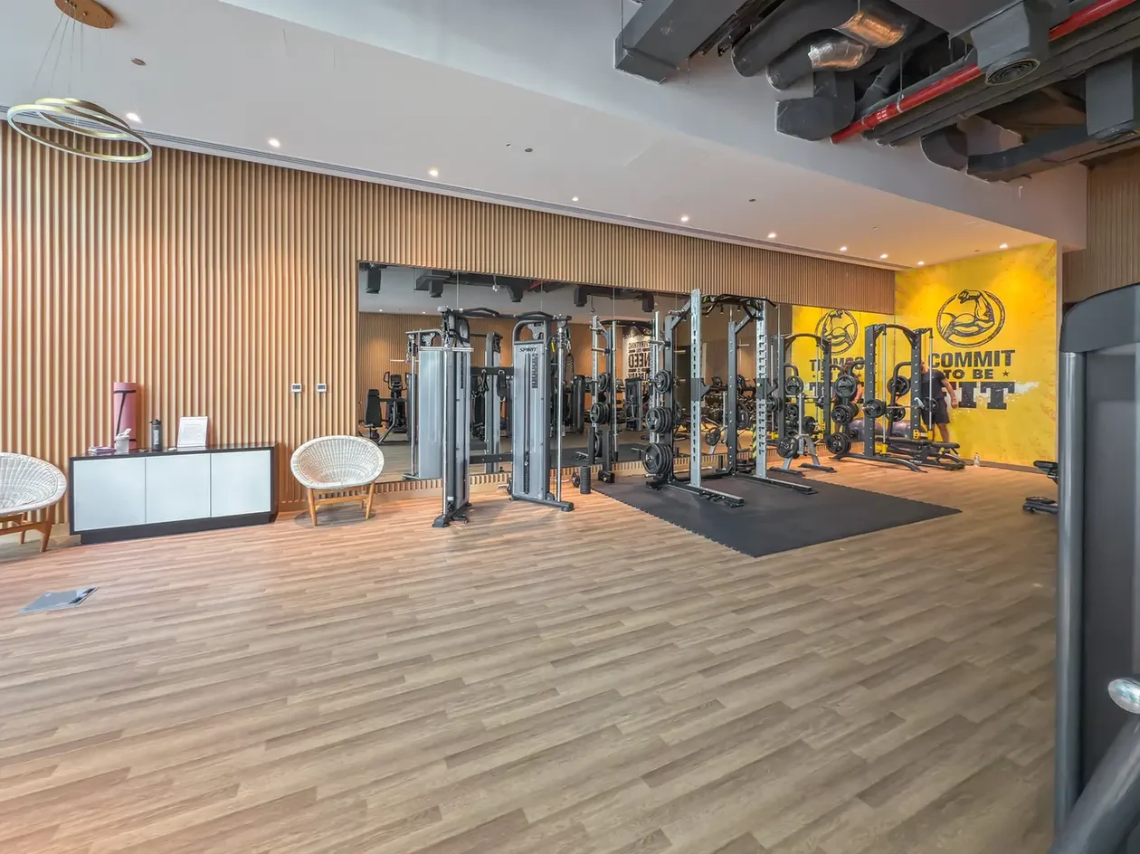 Open-plan gym with squat racks and free weights on a wood floor, a yellow mural reading 'Commit to be fit' in the corner.