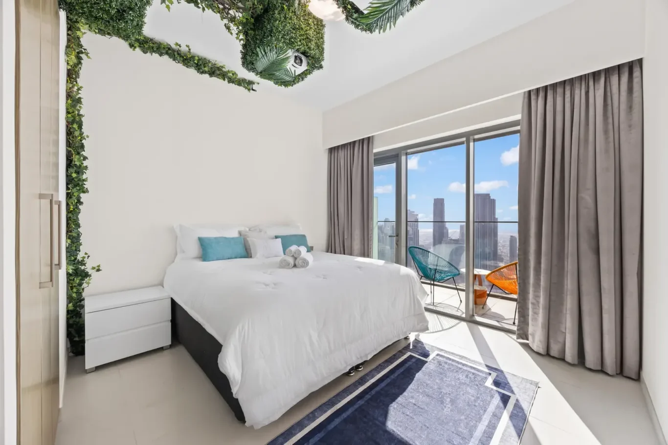 Bright, modern bedroom with white bed, teal pillows, and rolled towels; floor-to-ceiling windows open to a city skyline.
