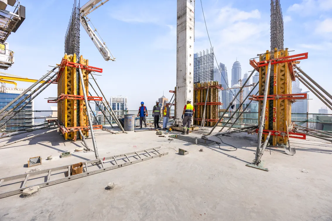 Construction workers on a rooftop with formwork and rebar columns under a crane rise, city skyline in the background