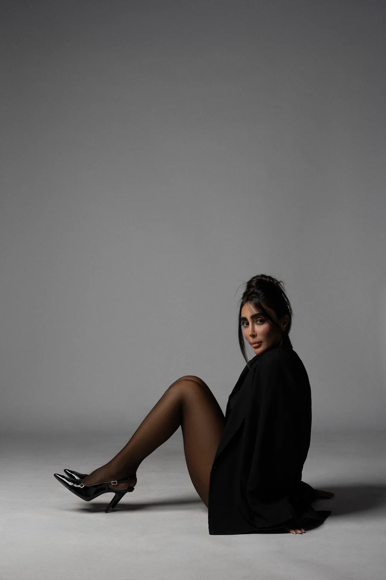 Woman seated on the floor in a black coat, wearing sheer tights and black high heels, posed against a gray studio backdrop.