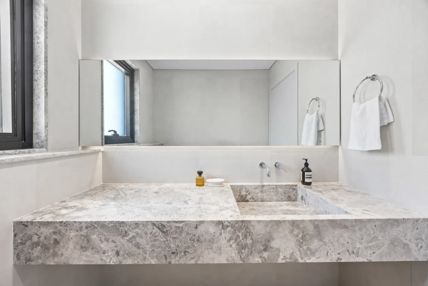 Modern bathroom vanity with a marble countertop, built-in sink, and large wall mirror; chrome towel rings hold white towels to the right.