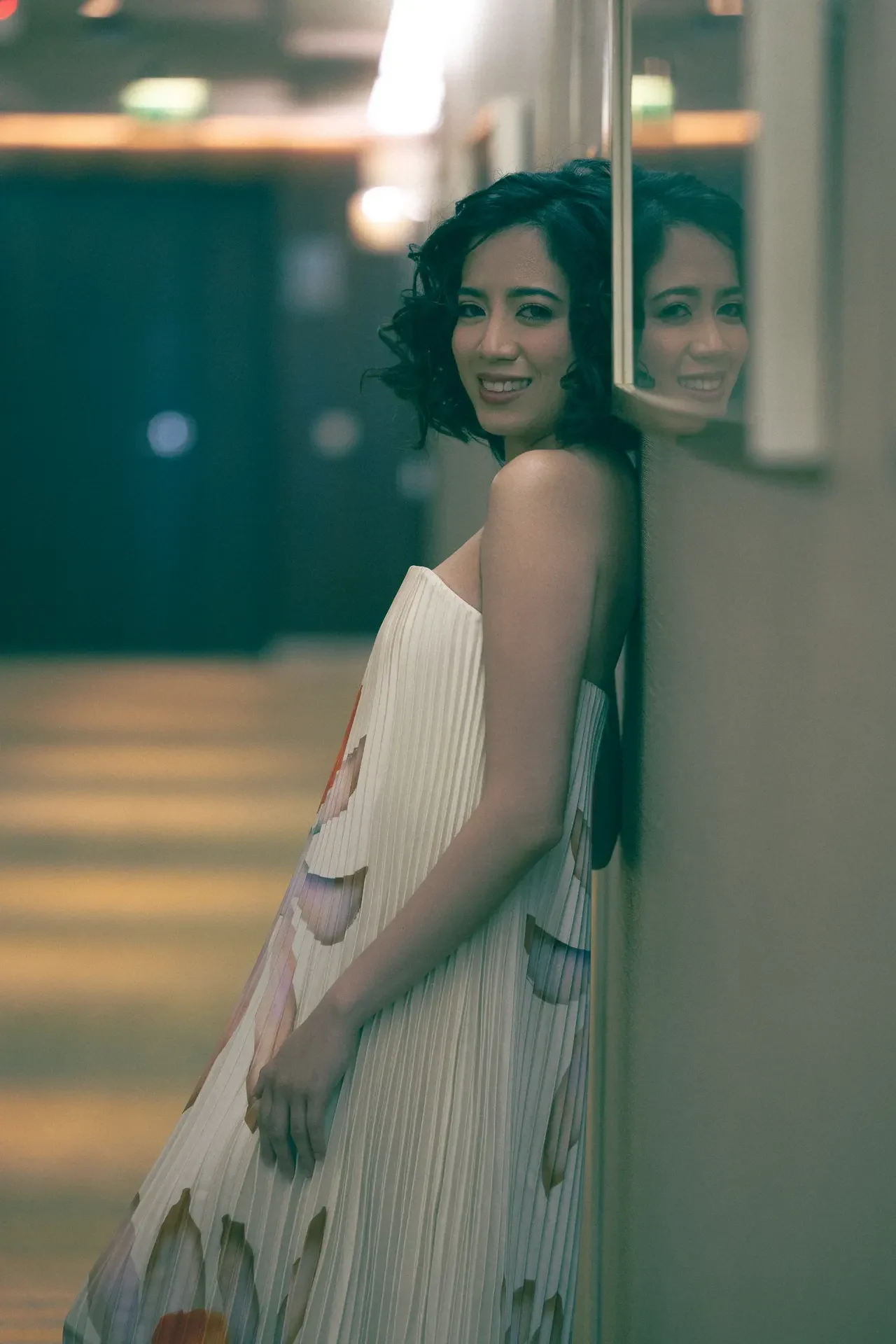Smiling woman in a light striped dress leaning against a glass wall, her reflection visible in the pane.