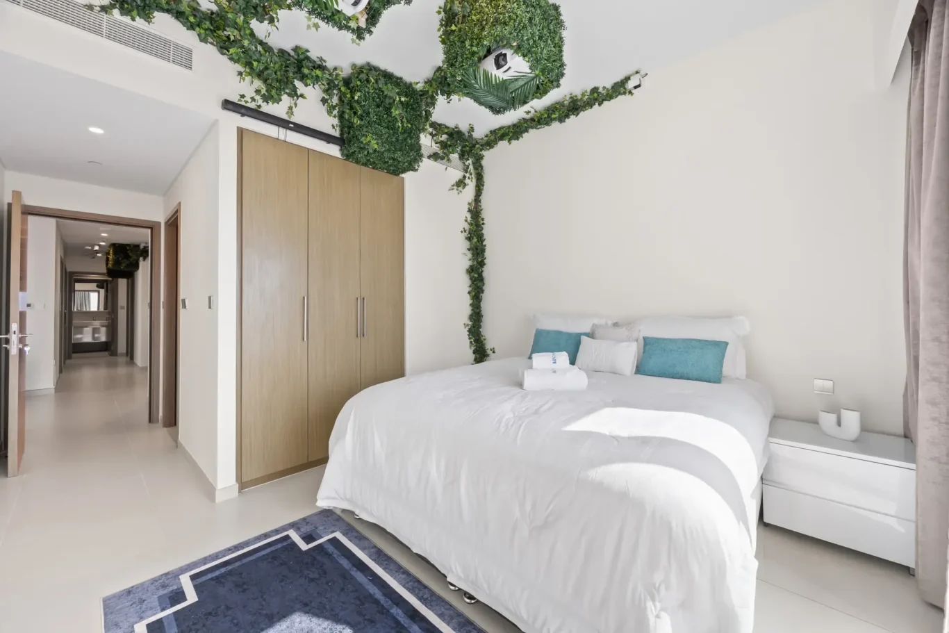 Bright bedroom with white bedding, blue pillows, and green trailing plants along the ceiling and wall, plus a wooden wardrobe and hallway view leftfully visible.