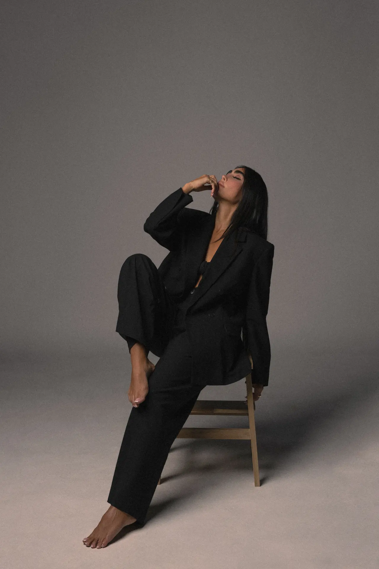 Woman in a black suit, barefoot, sits on a chair with one leg raised and head tilted back in a studio setting.
