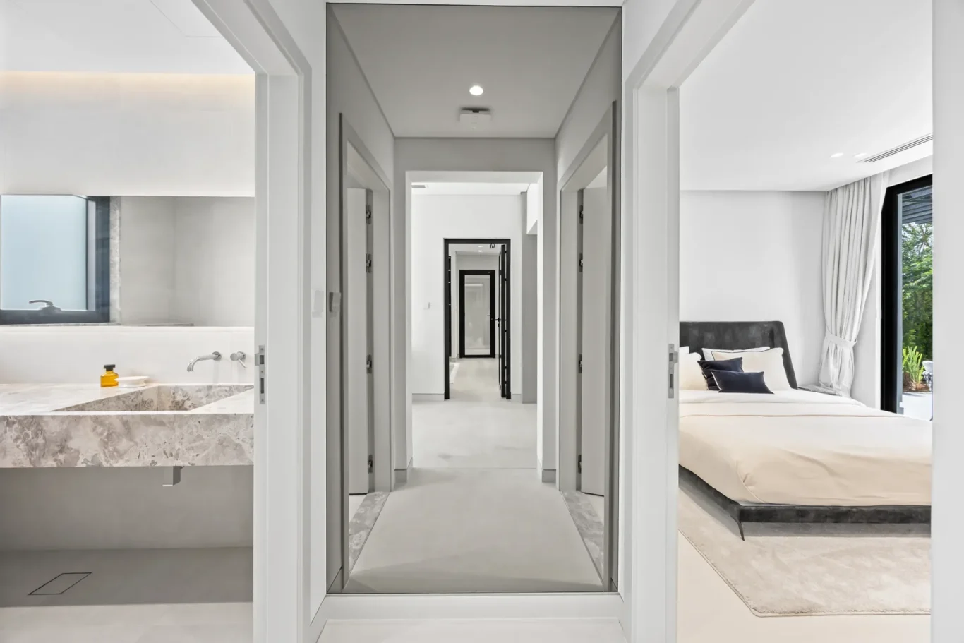 Long white hallway with open doorways, a marble bathroom on the left and a bedroom with a bed visible on the right.