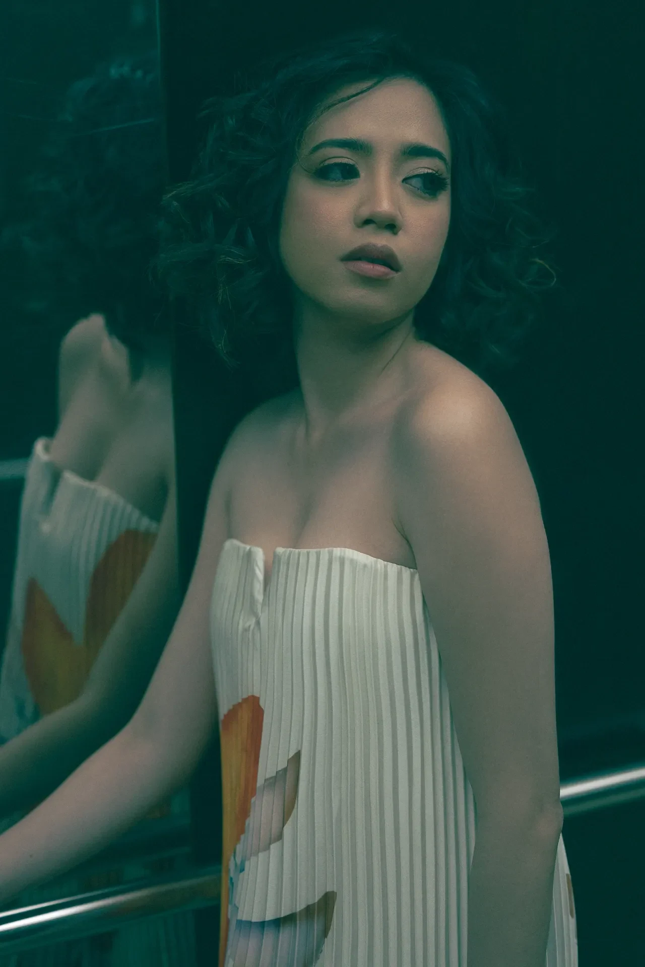 Young woman with short, wavy hair in a strapless white pleated dress standing near a dark reflective surface, looking off to the side.