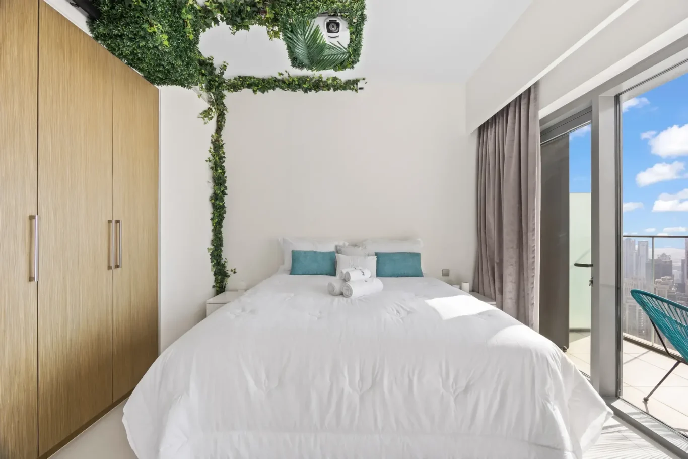 Modern hotel bedroom with white linen bed, teal cushions, wooden wardrobes, and ivy decor leading to a balcony with city view.",