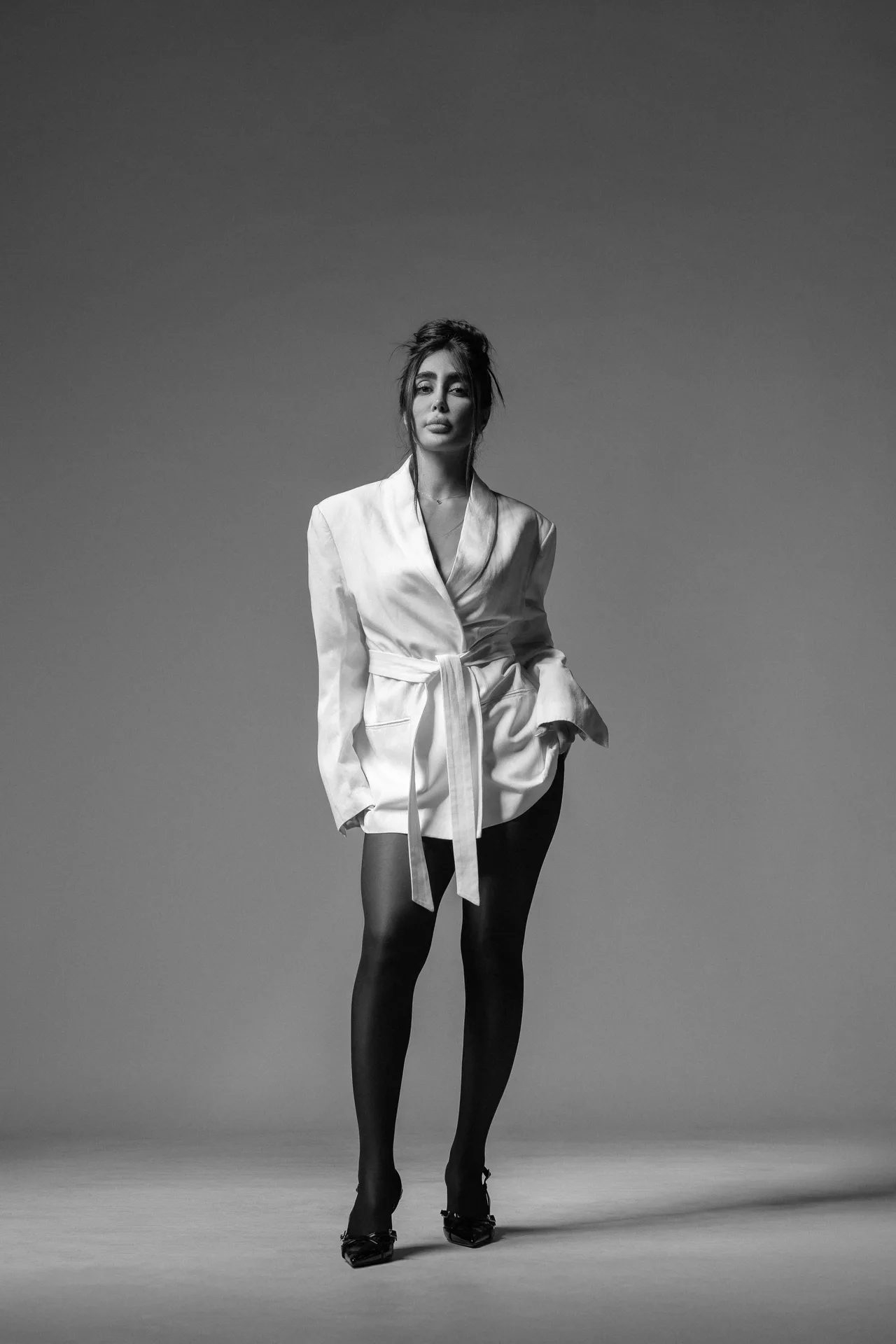 Monochrome fashion portrait of a woman standing in a white belted wrap blazer with dark tights and heels against a plain backdrop.