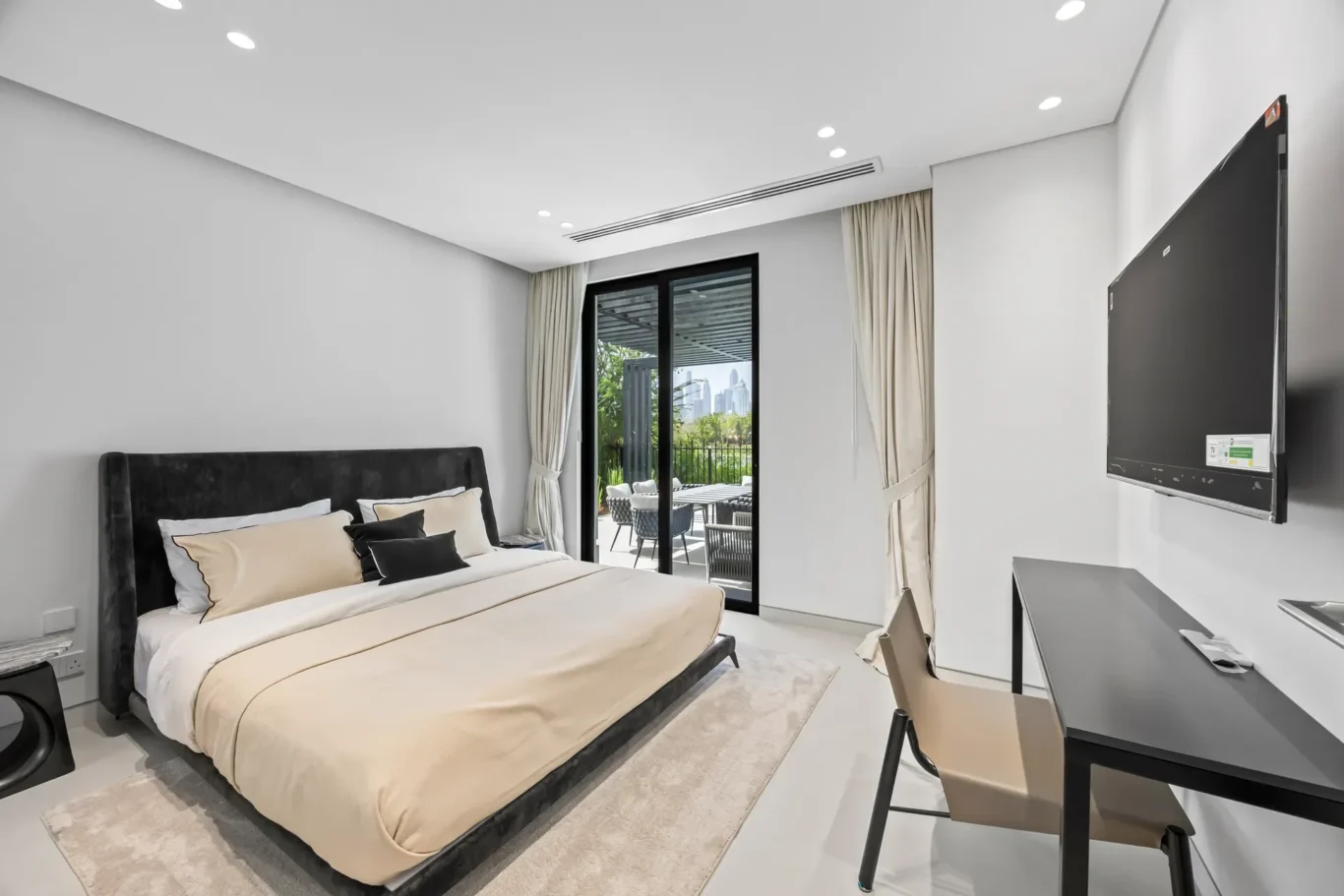 Modern bedroom with a black upholstered bed, cream bedding, and a rug; glass balcony doors with beige curtains open to an outdoor seating area.