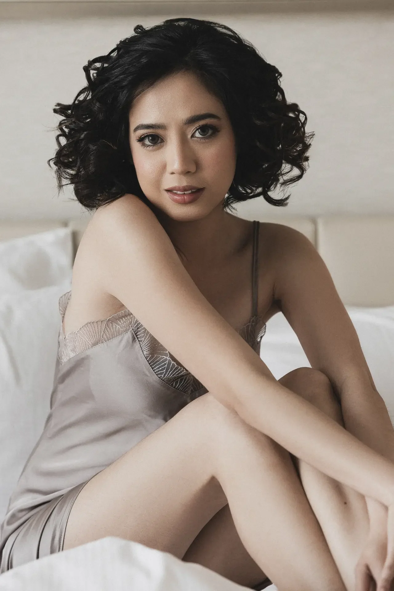 Woman with short curly black hair seated on a bed in a satin beige nightgown, gazing softly at the camera.