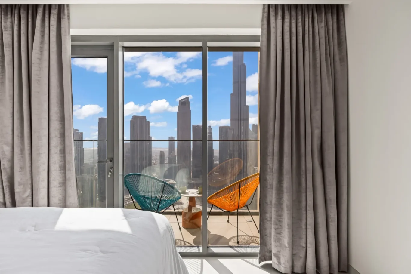Bedroom view with glass doors to balcony; teal and orange chairs and a small table outside against a city skyline and blue sky with clouds.