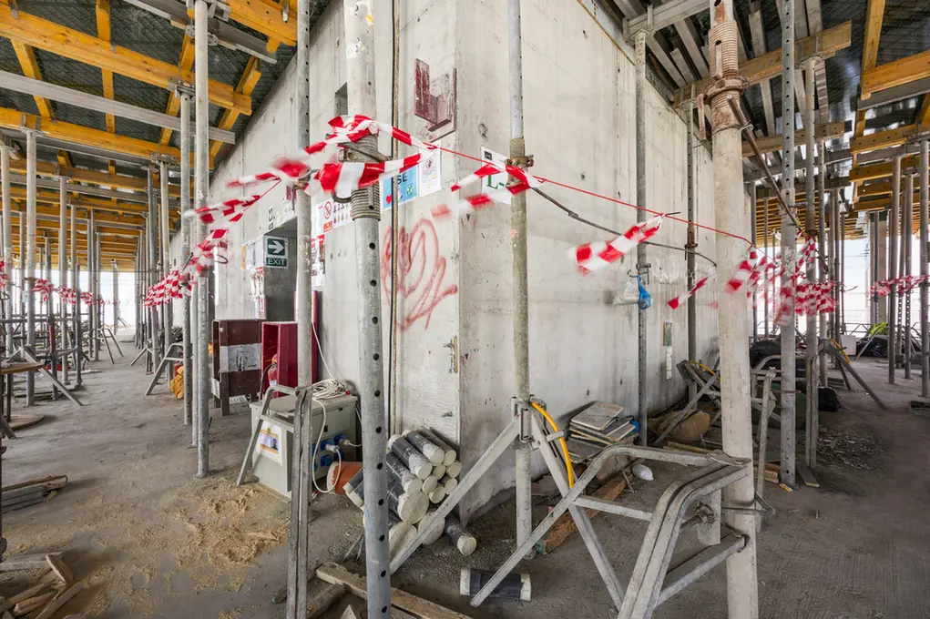 Construction site interior with metal scaffolding, concrete wall, and red-white safety tape across the area; clutter and building materials scattered on the floor.