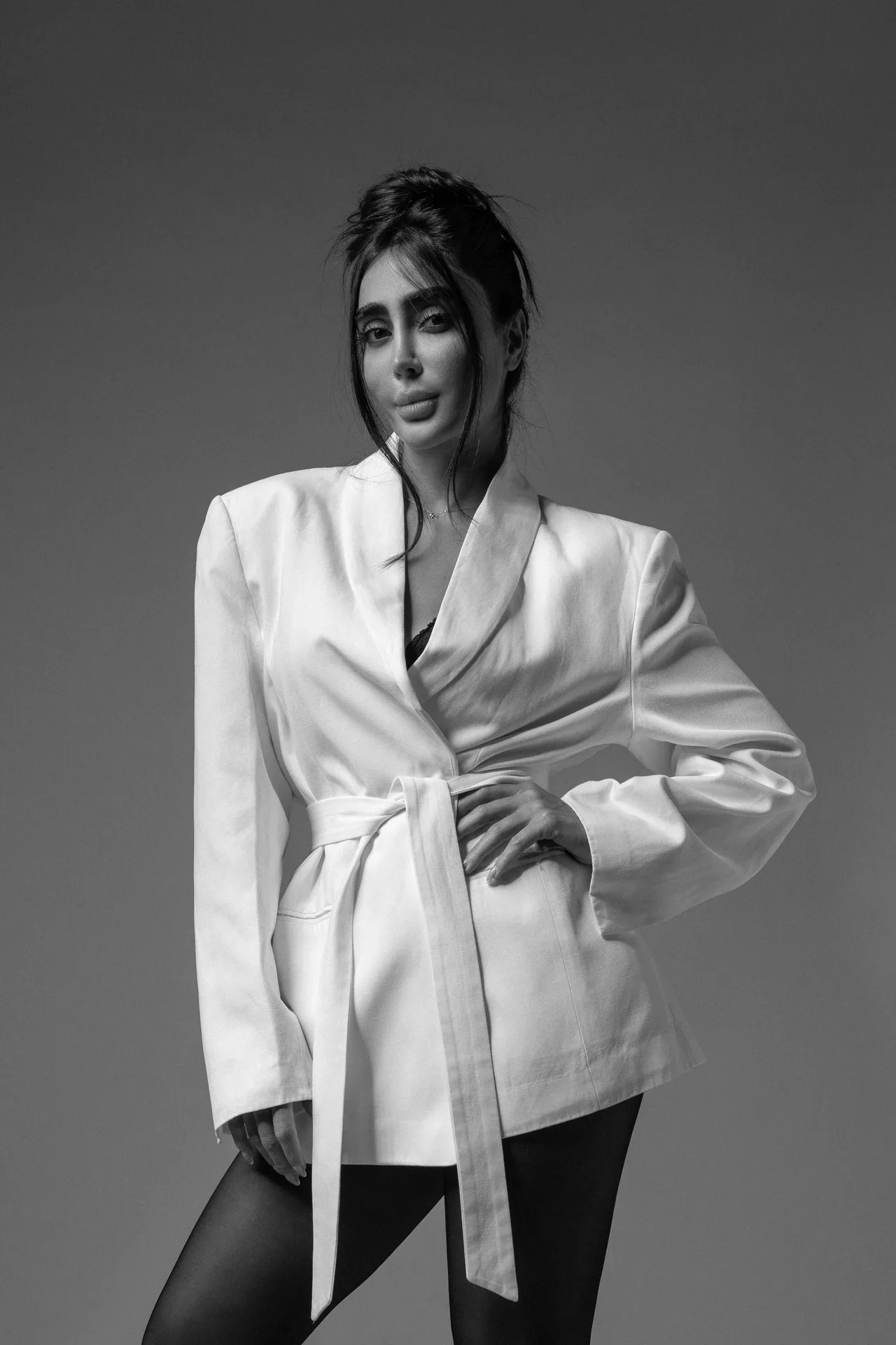 Stylish portrait of a woman in a belted white blazer, standing with hands on her waist against a gray studio backdrop, in black-and-white.