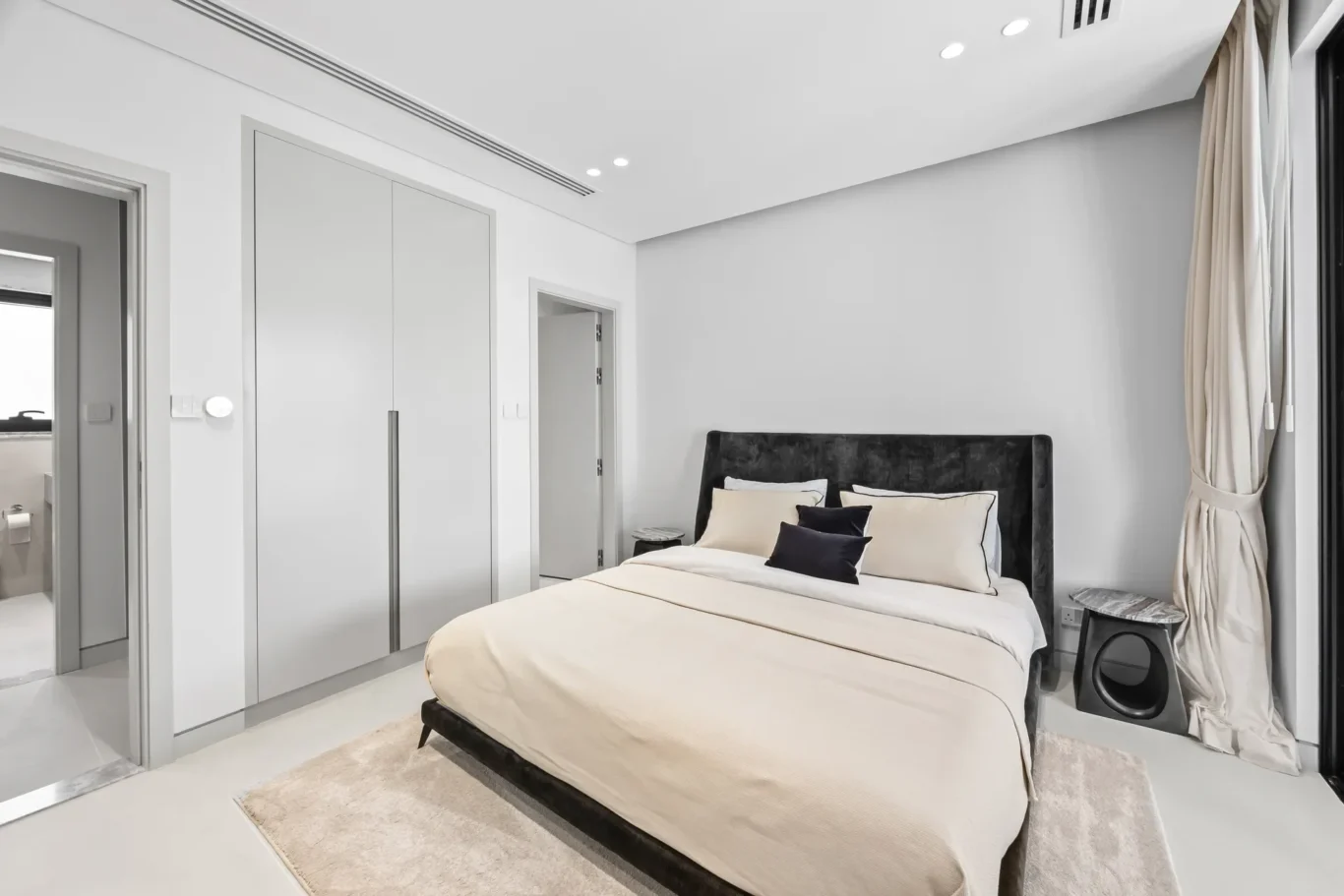 Modern bedroom with a large upholstered gray headboard bed, beige bedding, and multiple decorative pillows, on a light rug in a neutral-toned room.