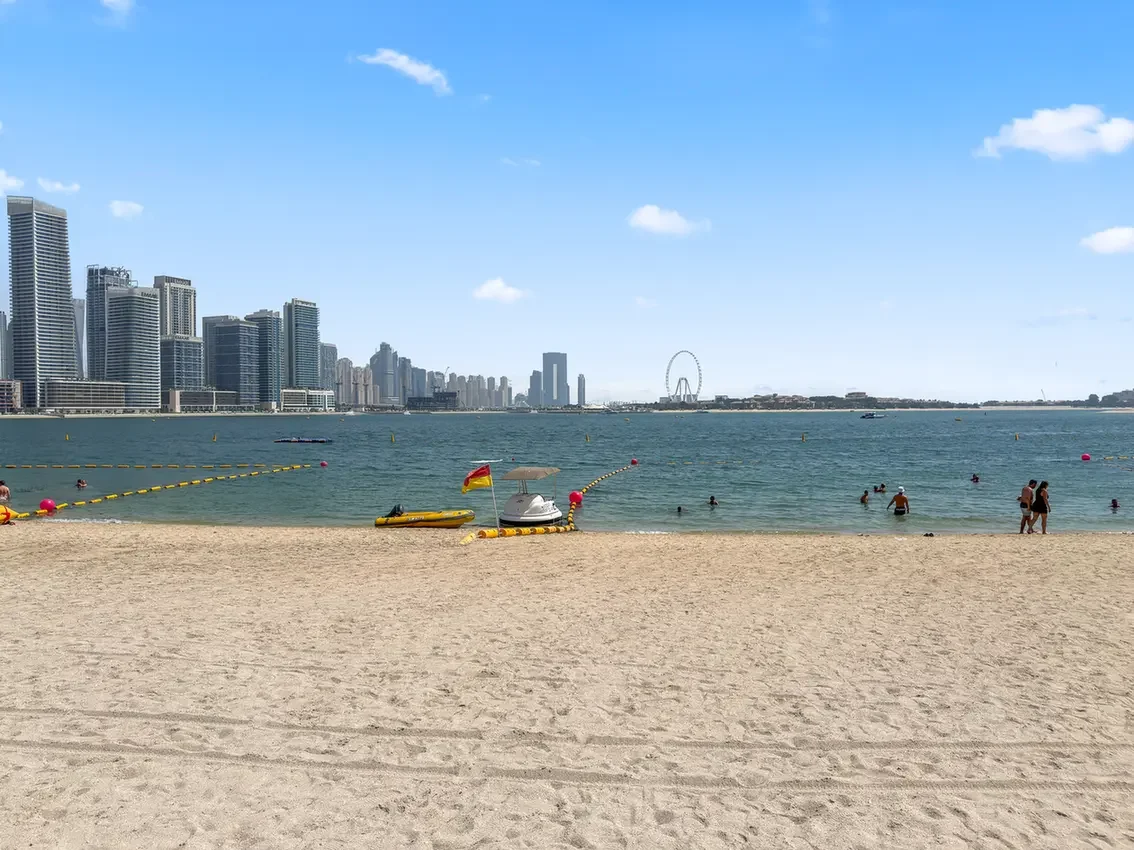 City skyline over a calm sea from a sandy beach; people swim and walk near the shore under a bright blue sky.