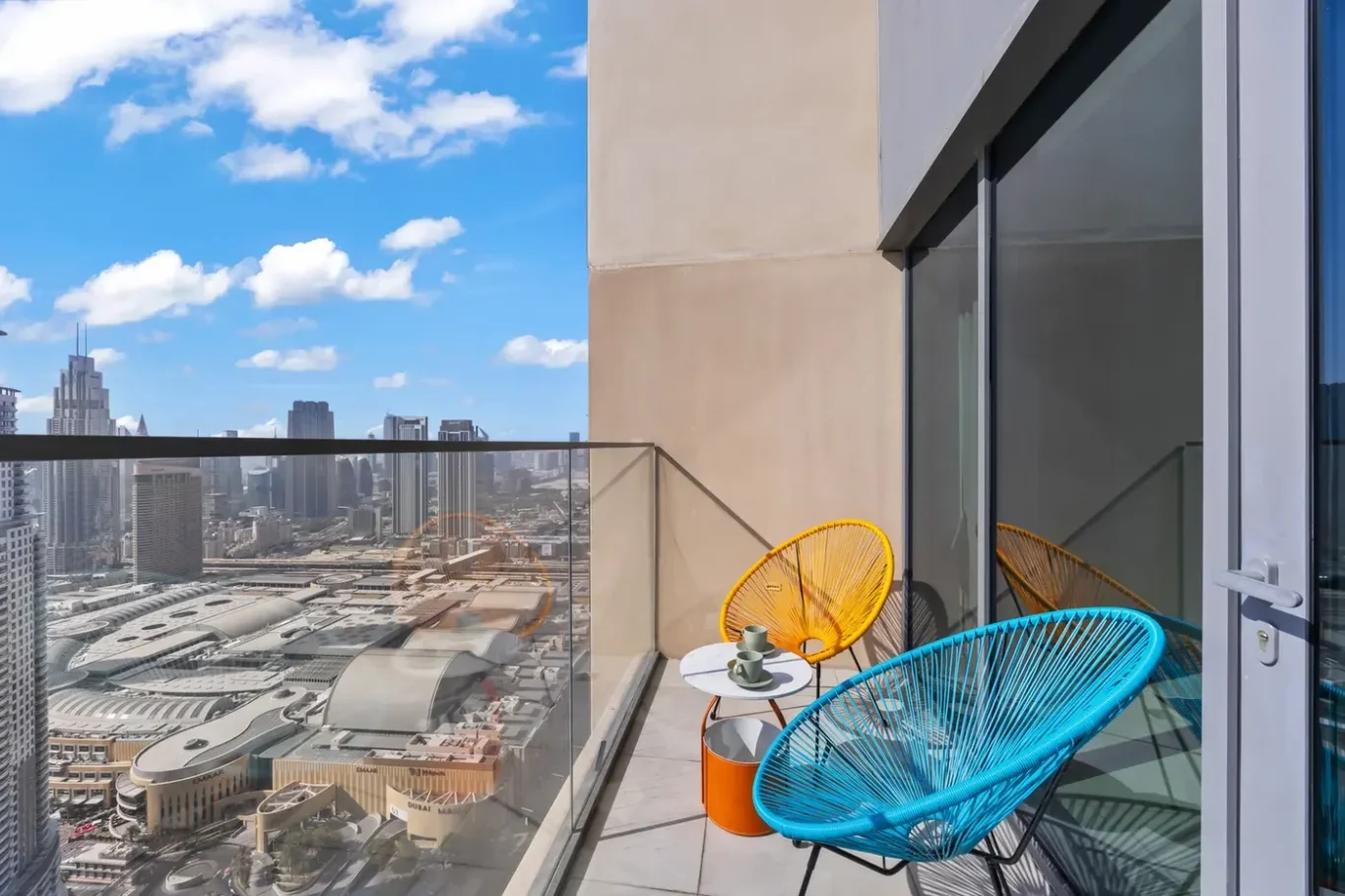 Colorful Acapulco chairs (blue and orange) on a balcony with a small table and cups, overlooking a city skyline and glass railing under a blue sky.