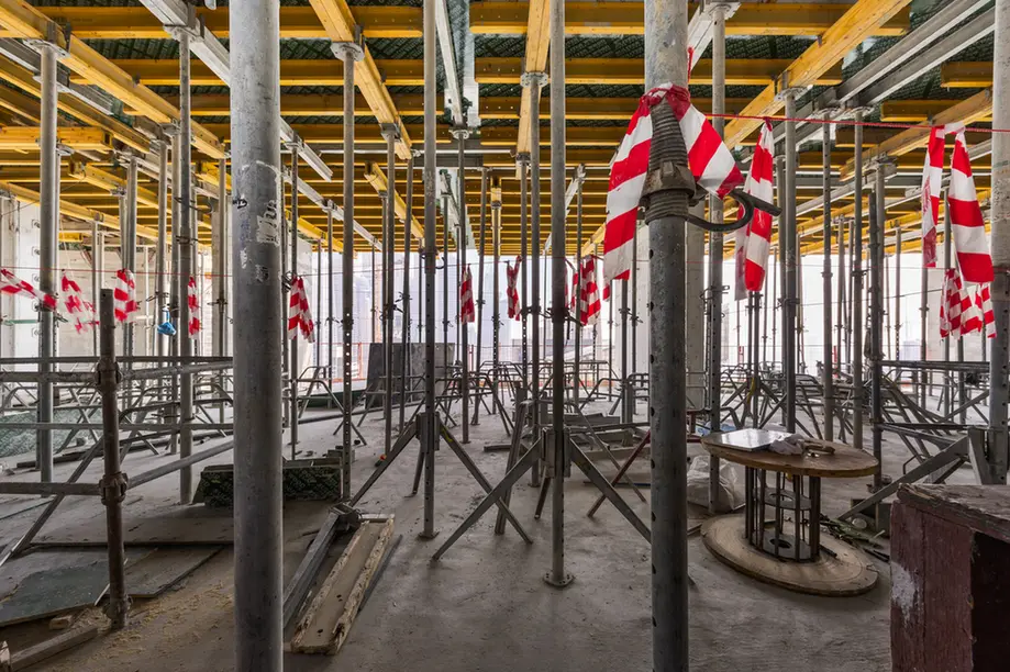 Interior of a construction site with numerous metal scaffolding poles and crossbars, yellow beams overhead and red-and-white safety tape tied to some poles.