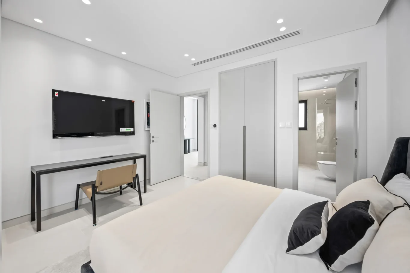 Minimalist hotel-style bedroom with a wall-mounted TV, desk and chair, seen from the bed, and open doors to a bathroom and hallway