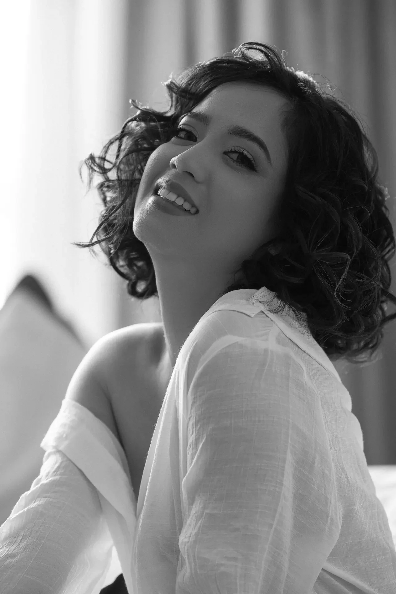 Black-and-white portrait of a smiling woman with short, wavy hair wearing a light blouse, head slightly tilted forward in a soft indoor setting.