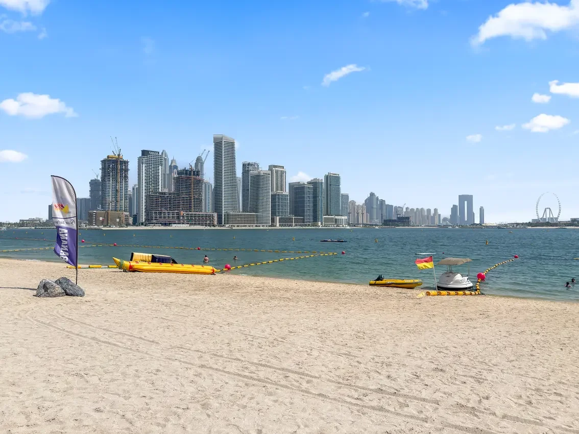 Sunny beach with yellow paddle boats near the shore and a city skyline across the water.