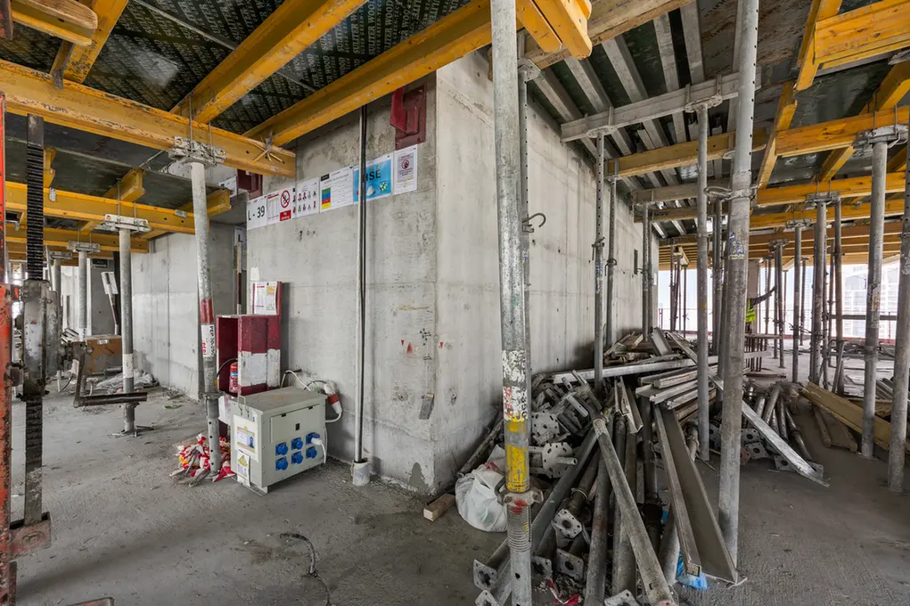 Construction site interior with concrete walls, yellow formwork beams, and scattered metal scaffolding and debris on a dusty floor.
