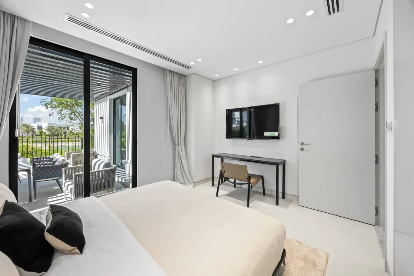 Bright modern hotel room with a bed in the foreground, desk and chair, wall-mounted TV, and glass doors opening to a balcony with outdoor seating.