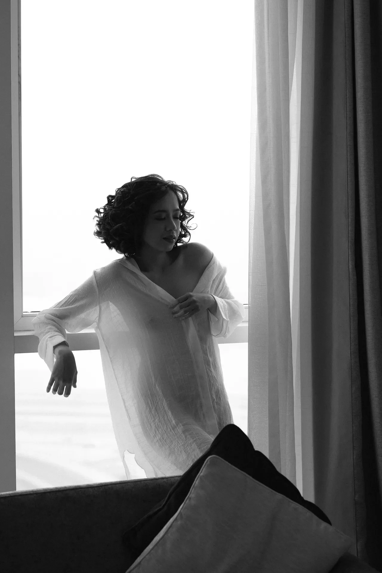 Woman with curly hair wearing a loose white off‑the‑shoulder dress standing by a window with sheer curtains, looking downward.