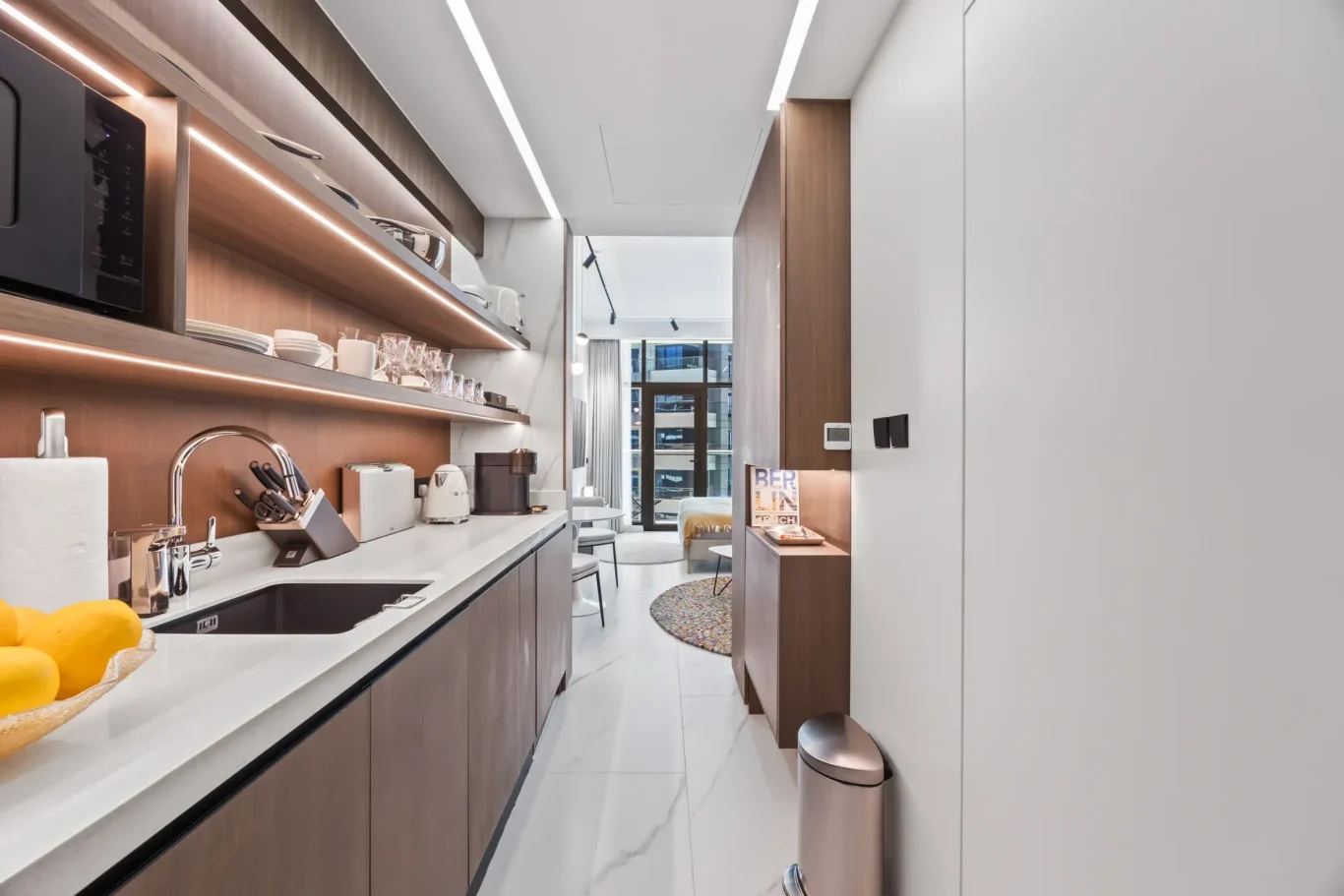 Modern galley kitchen with wood cabinets, white countertops, and open shelves displaying dishes and glassware.