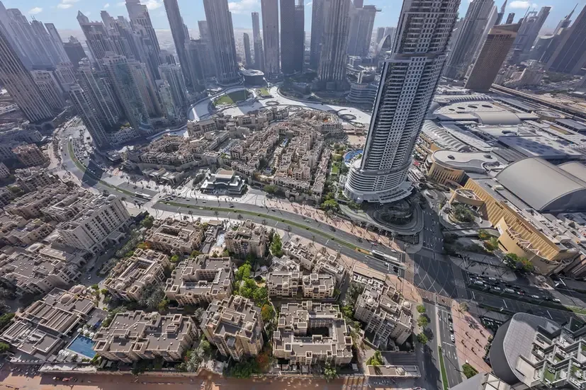 Aerial view of a modern cityscape with tall skyscrapers surrounding a circular road interchange and a cluster of low-rise beige buildings at the center. A curved high-rise tower rises above the surrounding area on the right. Parked roads and a large shopping/entertainment complex are visible nearby.