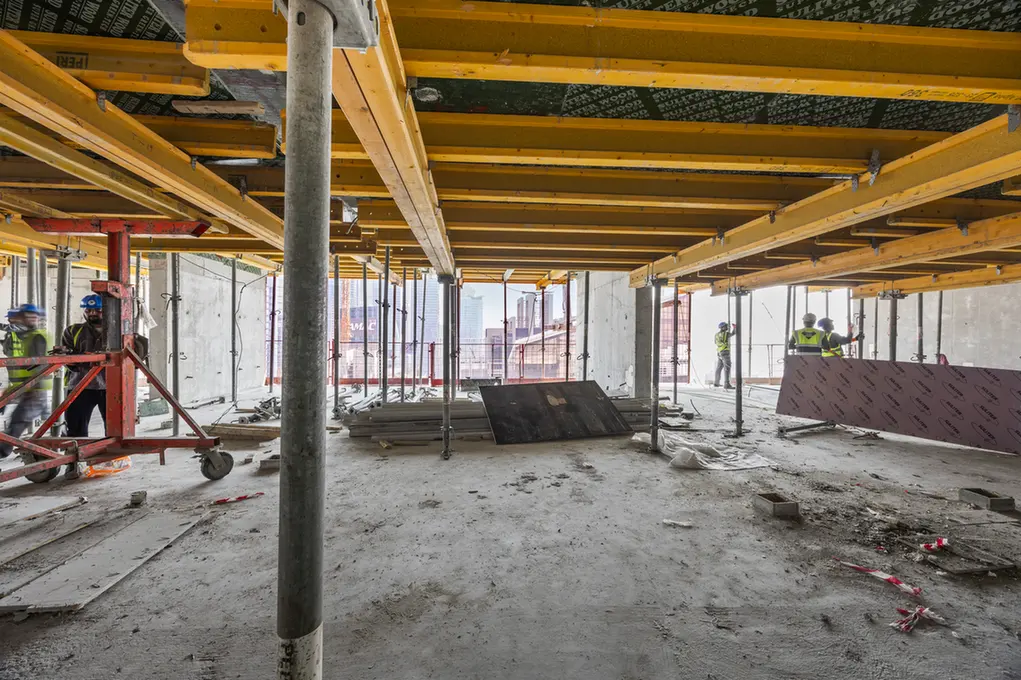 Construction site interior with yellow overhead beams, scaffolding, and workers in safety vests and hard hats.