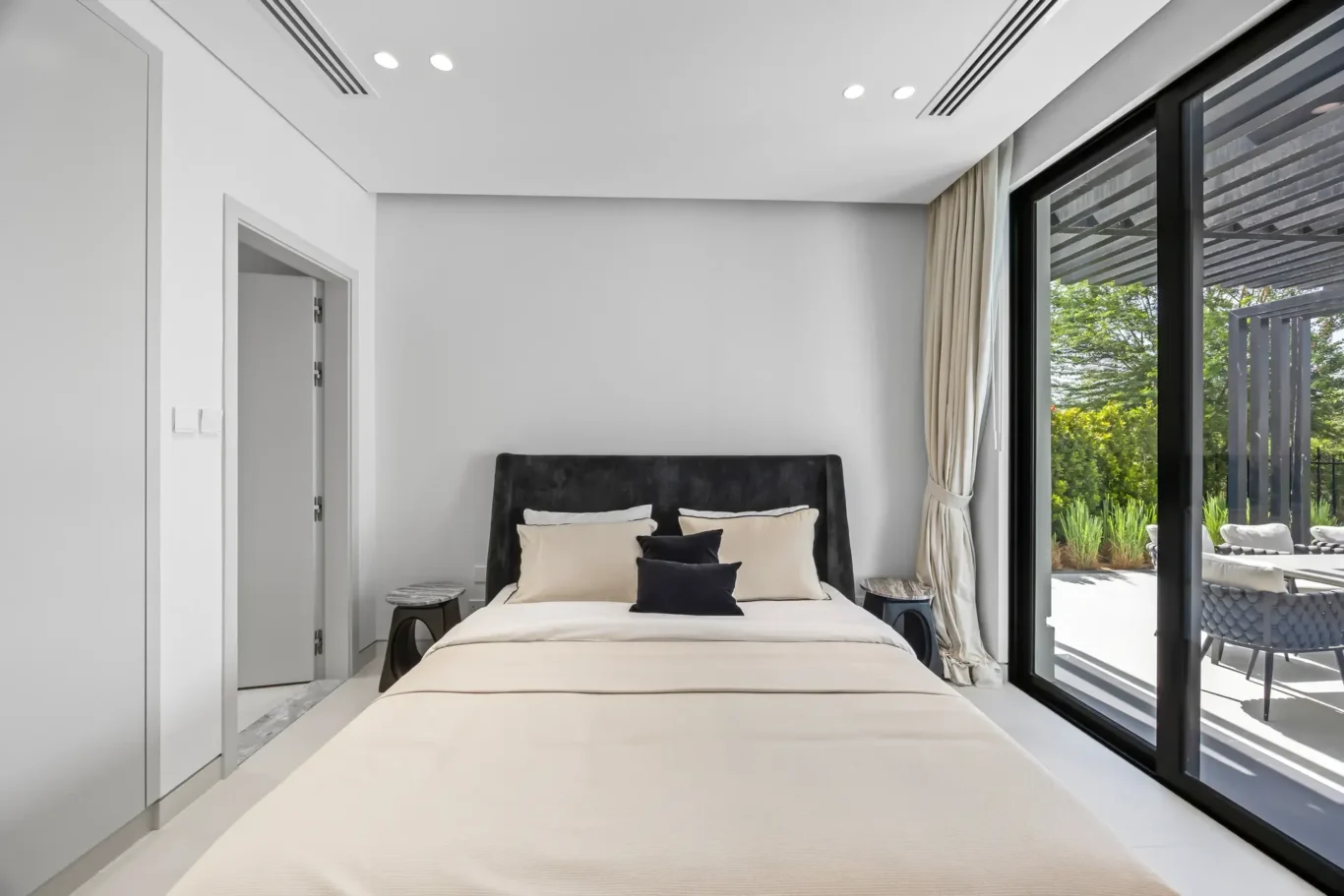 Bedroom with beige bedding and black headboard, two round nightstands, and large glass doors opening to a sunny patio.