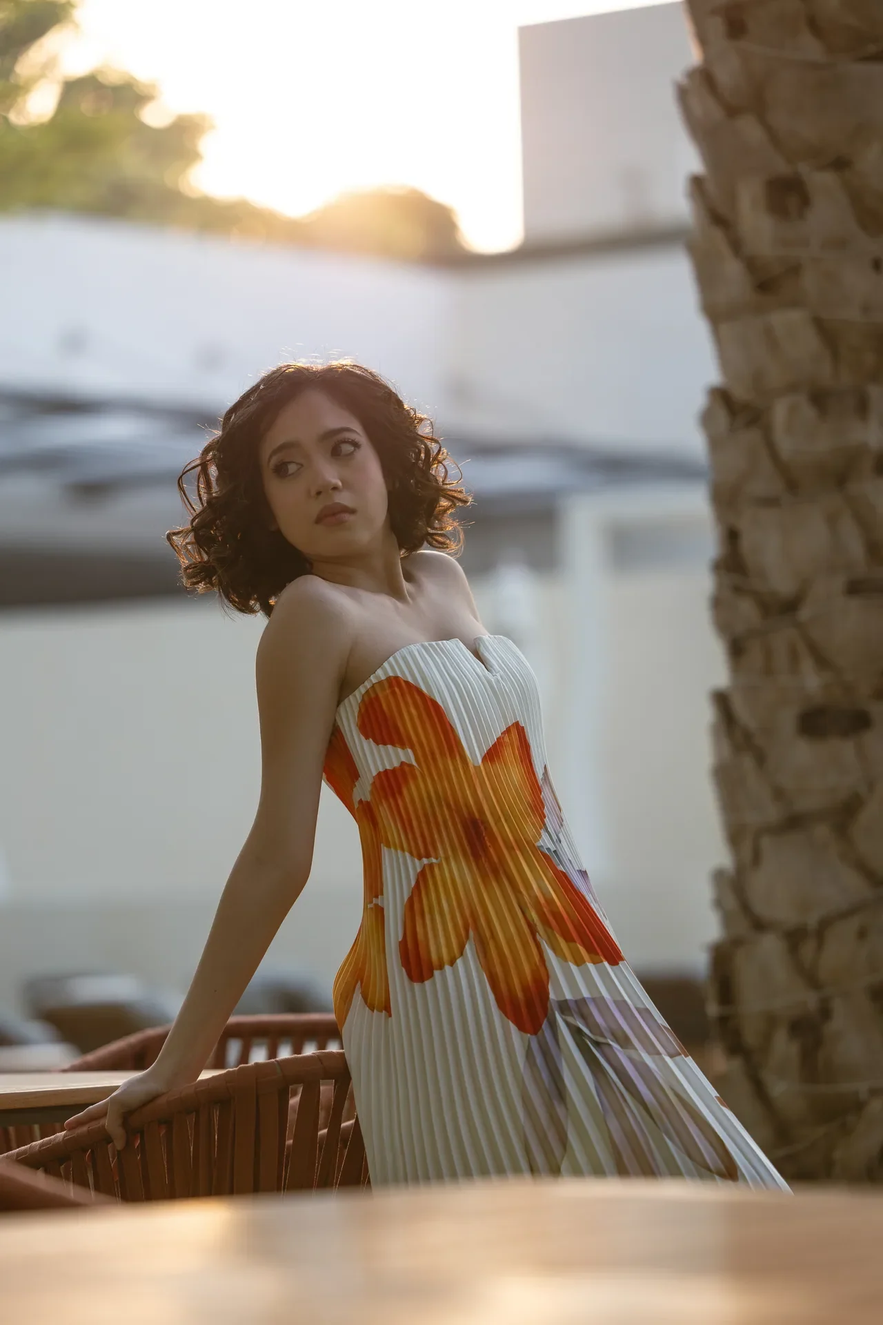 Woman with curly hair in a white pleated dress with orange floral pattern leaning on a wooden railing at sunset.
