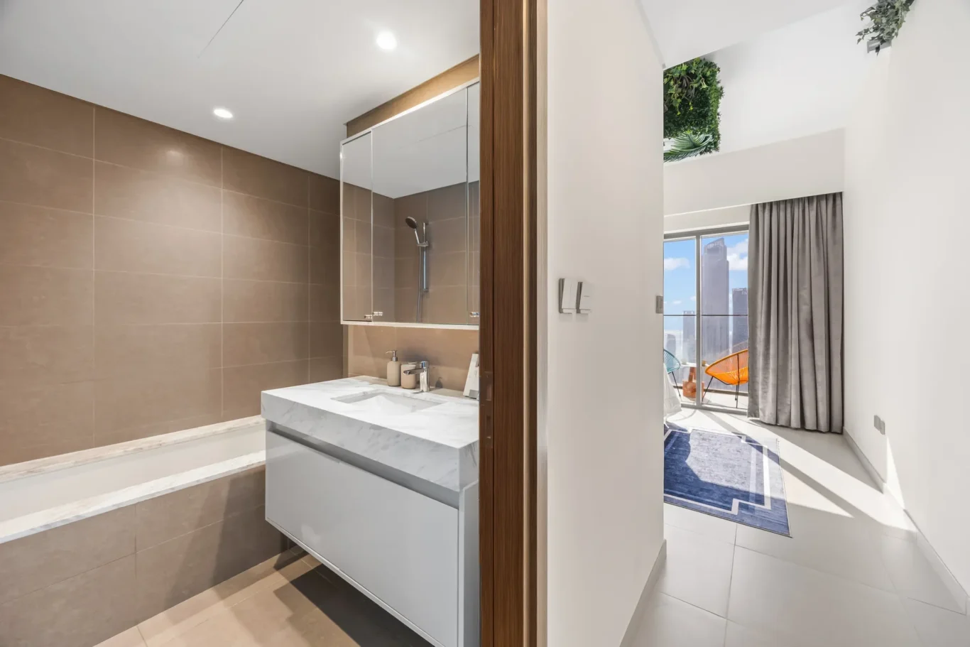 Modern bathroom with brown tiled walls, white marble vanity, and glass-front shower; wooden door frame opens to a bright living area.