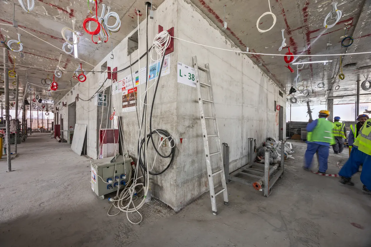 Construction site interior with exposed wiring, ladders, and workers in hi-vis vests around a concrete wall.